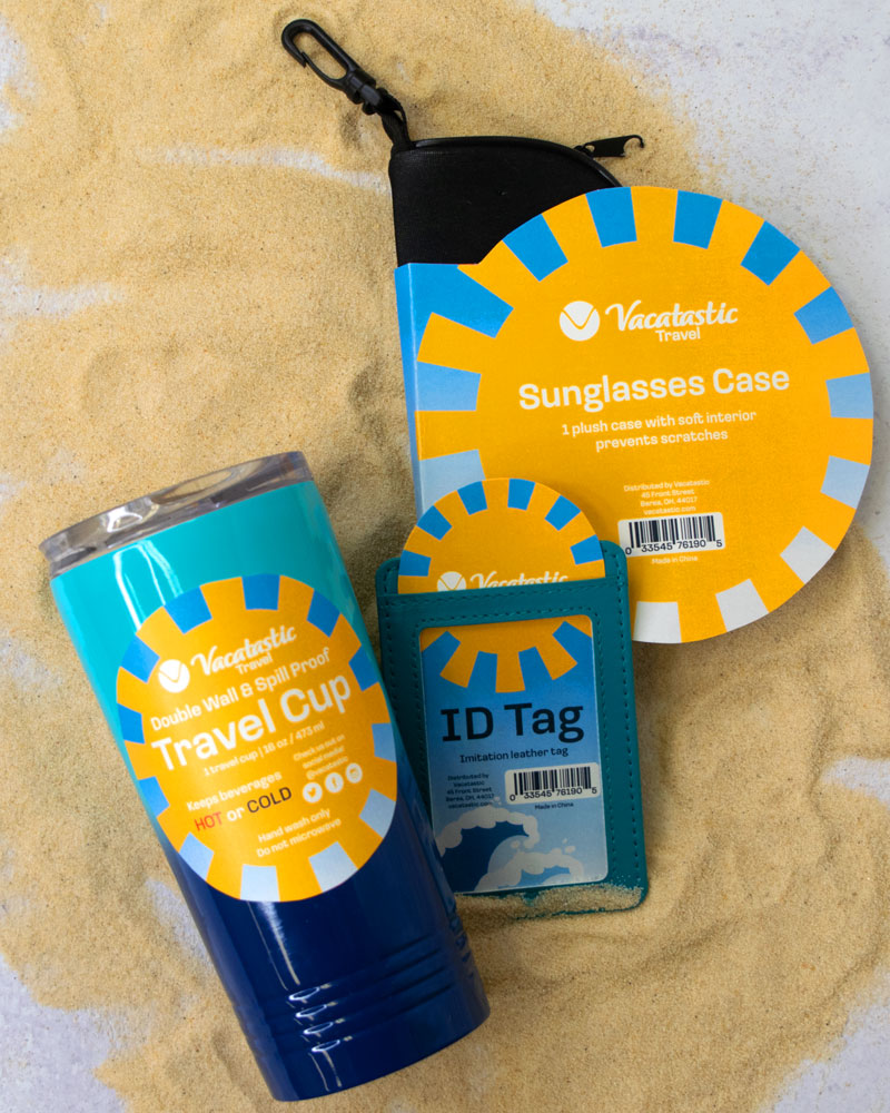 Travel cup, ID tag, and sunglasses case with Vacatastic packaging laying on sand 